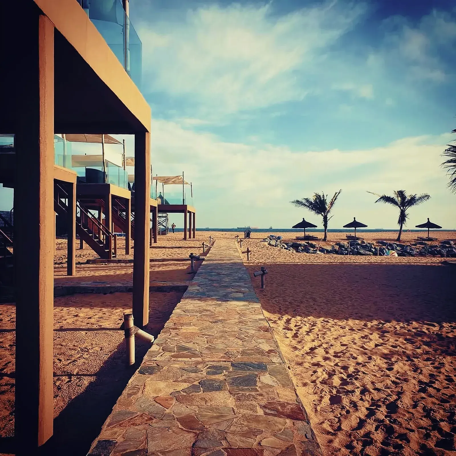Serene beach scene with a stone walkway, palm tree, and beach umbrella