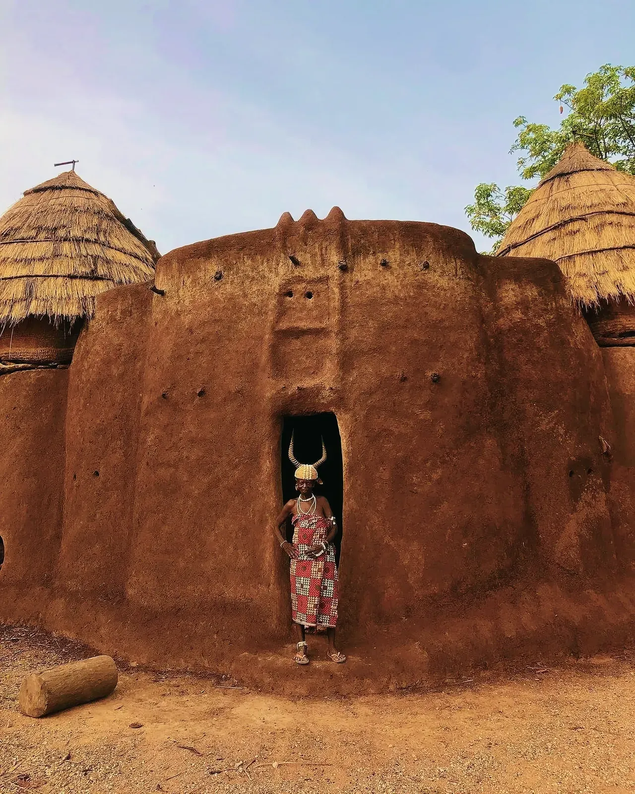 Woman in a red and white dress standing in front of a rustic mud house in Togo